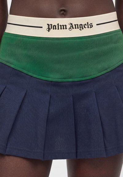 Ζώνη μέσης με το λογότυπο "Palm Angels" σε εκρού ταινία πάνω από πράσινο επικάλυμμα και σκούρο μπλε πλισέ φούστα σε άτομο με σκούρο δέρμα.
