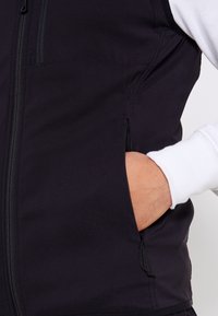 Händchen in der Tasche einer schwarzen Zip-up-Weste, die über einem weißen Langarmhemd getragen wird, Nahaufnahme, die den Stofftextur und die Reißverschlussdetails zeigt.