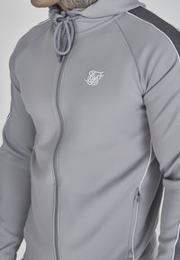 Grå zip-up hoodie med en texturerad logga, dragsko i huvan och kontrasterande svarta axelsträck med vita detaljer.