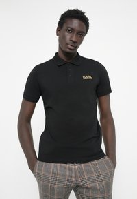 Fekete pólóing puha anyagból, gallérral, az előnyak bal oldalán arany "KARL LAGERFELD" logóval. Kockás nadrággal viselve.