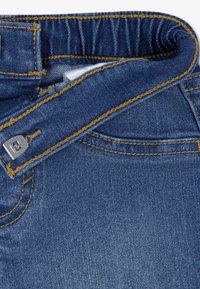 Μπλε τζιν πουκάμισο με καμπύλη μέση, ραφές σε κίτρινο χρώμα και μεταλλικό κουμπί. Το ύφασμ α έχει λεία υφή.