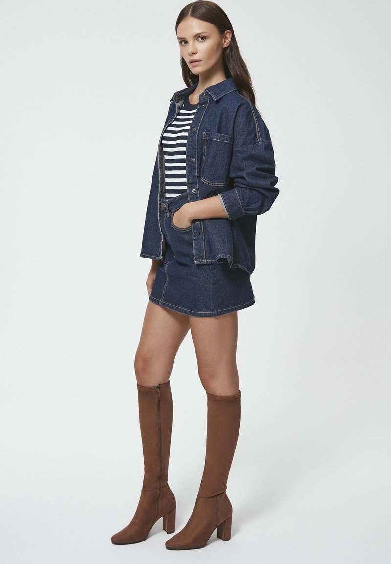 Jeansowa kurtka o luźnym kroju, paskowana koszula z długim rękawem, jeansowa spódnica oraz brązowe zamszowe kozaki za kolano na słupkowym obcasie.