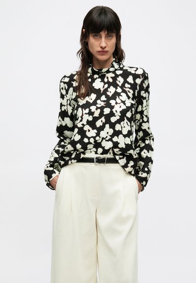 Melna blūze ar gaišiem ziedu rakstiem, apaļu apkakli, garām piedurknēm un brīvu piegriezumu; saskaņota ar krēmkrāsas biksēm ar augstu jostasvietu un melnu jostu.