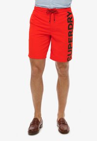Bañador rojo con cintura ajustable y un gran logo negro "SUPERDRY" en la pierna izquierda. Tela suave con un ajuste regular.