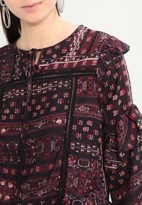 Blusa nera floreale con maniche arricciate, caratterizzata da intricati motivi rossi e bianchi, dettagli in pizzo e scollatura a keyhole.