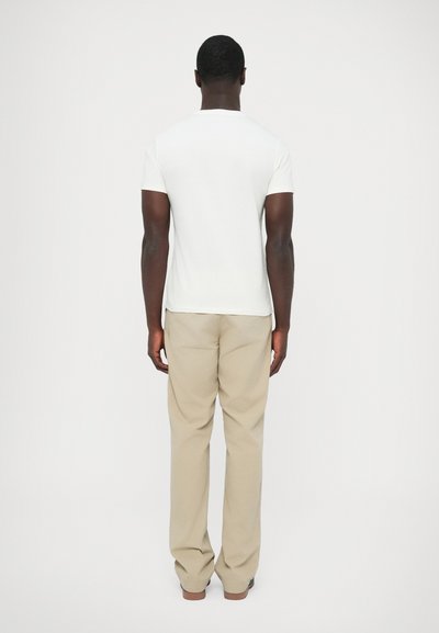 T-shirt branca de mangas curtas com gola redonda, combinada com calças bege claras, vista por trás, mostrando um design simples e forma ajustada.