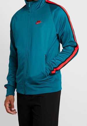 Mężczyzna ubrany w turkusową kurtkę na zamek błyskawiczny z czerwonymi i czarnymi paskami na rękawach oraz czerwonym logo Nike na piersi, czarne spodnie, ręka w kieszeni.
