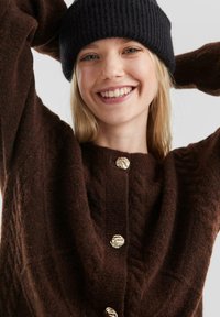 Bruine gebreide cardigan met een textuurpatroon, voorzien van een ronde halslijn en gouden knopen. Draag het met een zwarte geribbelde beanie.