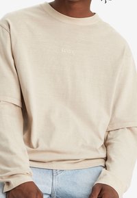 Osoba ubrana w beżową, długą koszulę z warstwami z logo "Levi's" na klatce piersiowej oraz jasnoniebieskie dżinsy, z rękami spoczywającymi w okolicy talii.