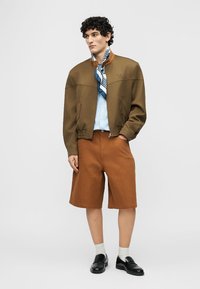 Homme portant une veste aviateur marron olive, une chemise bleu clair, une écharpe à motifs bleus, un short marron, des chaussettes blanches et des mocassins noirs.