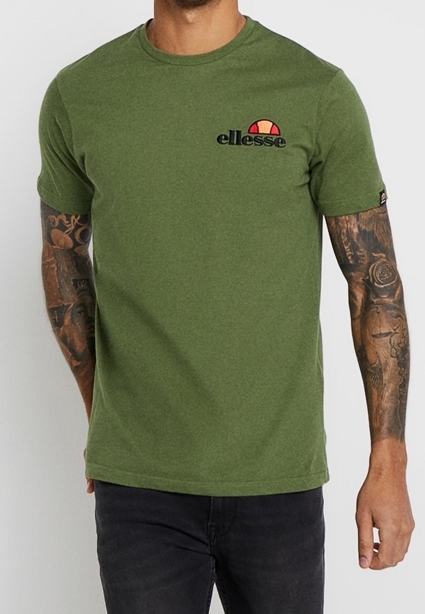 Homme portant un t-shirt ellesse vert olive avec un logo sur la poitrine gauche et un jean noir, montrant des bras tatoués sur un fond uni.