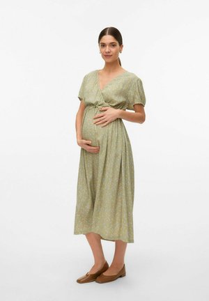Graviditetskjole i lys grøn stof med små blomsterprint, V-hals, korte puffærmer og en samlet talje. Båret med brune flade sko.