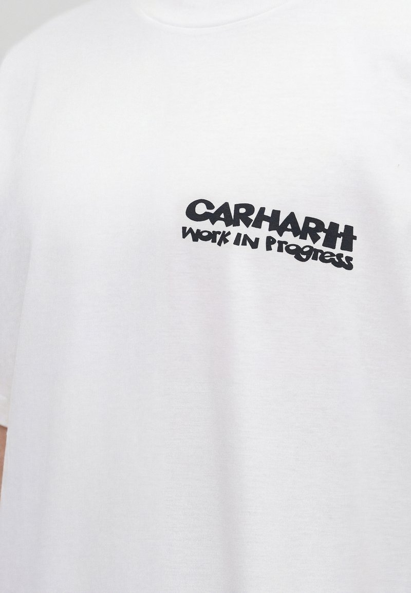 T-shirt bianca con testo nero "CARHARTT Work In Progress" stampato sulla parte superiore sinistra del petto.
