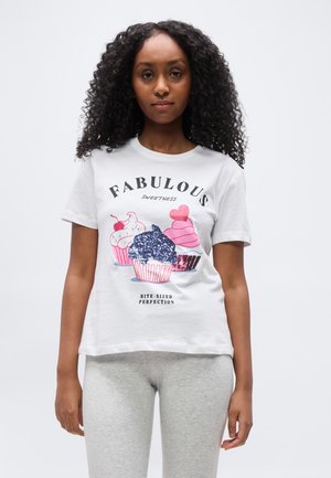 Kvinde med krøllet hår iført en hvid T-shirt med tre dekorerede cupcakes og ordene "FANTASTISK SØDME I BIDESSTØRRELSE PERFEKTION."