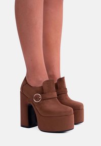 Scarpe platform in suede marrone con tacchi alti, punta tonda, cinturino con dettaglio di fibbia in metallo e un tacco spesso nero.