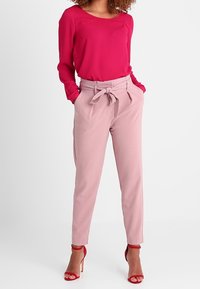 Blouse rose à manches longues, associée à un pantalon tailleur rose clair avec une taille à nouer, et des sandales à talons hauts rouges avec des lanières.