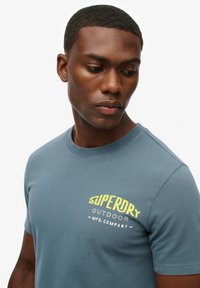 Camiseta de cuello redondo y manga corta en algodón azul oscuro, con un estampado gráfico amarillo en el lado izquierdo del pecho que dice "Superdry Outdoor."