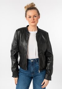 Zwarte leren bomberjack met een staande kraag, ritsluiting en gathering aan de manchetten. Gecombineerd met een wit T-shirt en hoge blauwe jeans.