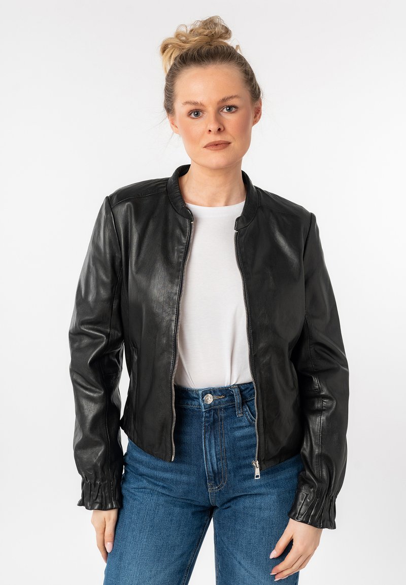 Zwarte leren bomberjack met een staande kraag, ritsluiting en gathering aan de manchetten. Gecombineerd met een wit T-shirt en hoge blauwe jeans.