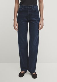 Jeans a gamba larga blu scuro in denim, caratterizzati da una vita alta, tasche anteriori e una texture pulita e liscia. Abbinati a un top nero.