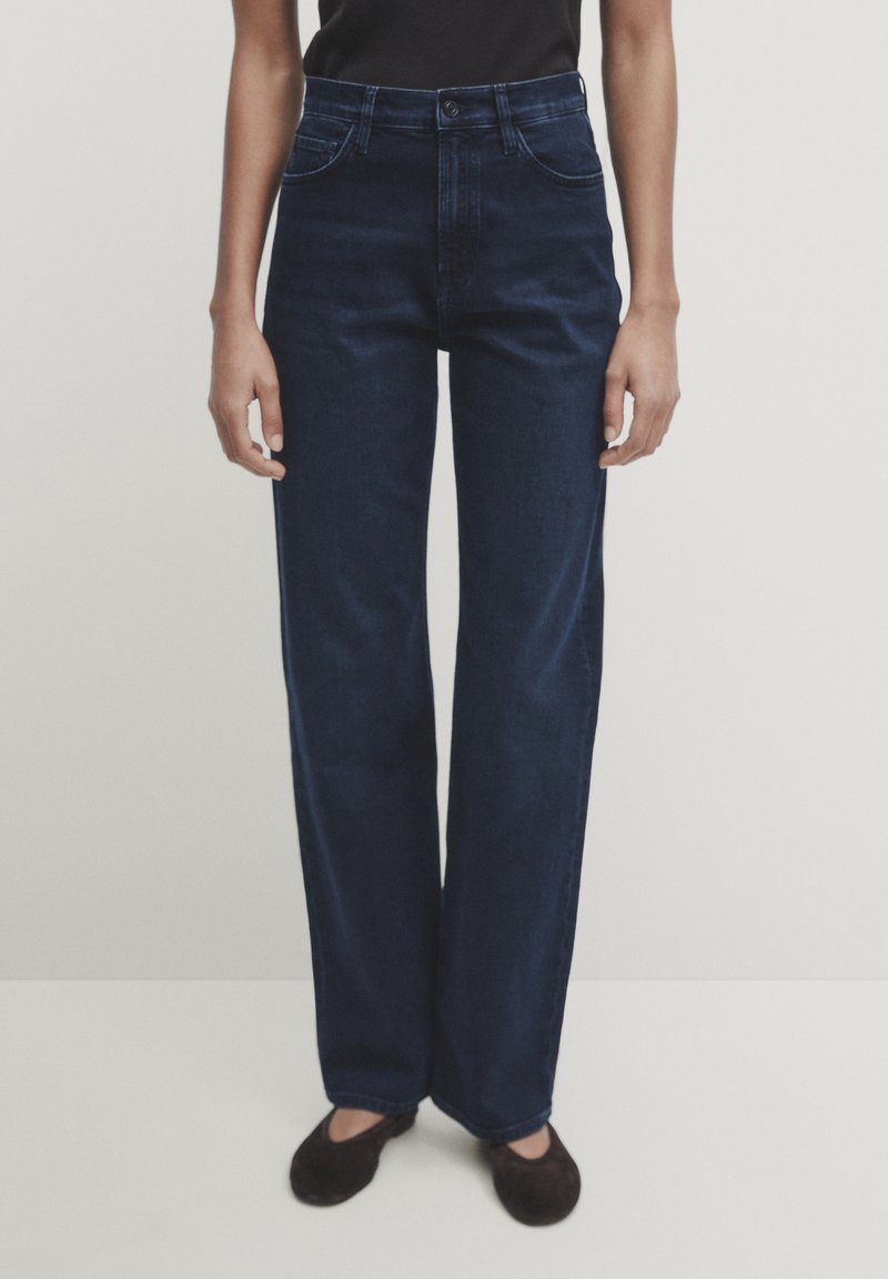 Jeans a gamba larga blu scuro in denim, caratterizzati da una vita alta, tasche anteriori e una texture pulita e liscia. Abbinati a un top nero.