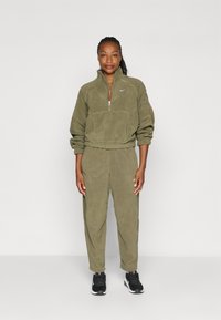 Sudadera de forro polar verde oliva con cremallera de un cuarto y puños elásticos, combinada con zapatillas deportivas negras con detalles en blanco.