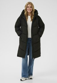 Sort puffershirt med hætte, der har lynlåslukning og lomme med klap. Sammen med en hvid sweater og blå flagrende jeans.
