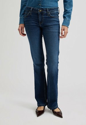 Femme portant un jean bootcut bleu foncé, une chemise en jean bleue et des talons pointus marron foncé, debout devant un fond clair.