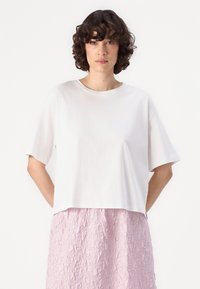 T-shirt blanc court à manches courtes, avec un col rond. Porté avec une jupe rose texturée, présentant une coupe décontractée et un tissu doux.