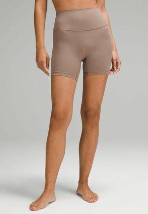Barfodet person iført beige, tætsiddende cykelshorts med høj talje, stående foran en ensartet lys grå baggrund.