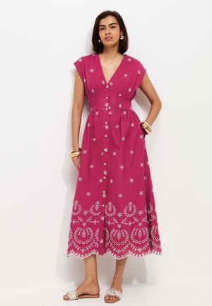 Kobieta w bezrękawnikowej sukience midi w kolorze magenta z białą kwiatową haftem i guzikami z przodu, połączona z białymi sandałami wsuwanymi.