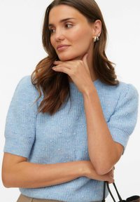 Pull à manches courtes tricoté bleu clair avec tissu texturé. Le modèle pose le menton sur la main, mettant en valeur les fibres douces et la coupe décontractée.