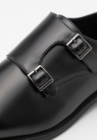 Chaussure en cuir noir avec une finition lisse, dotée de deux boucles en argent. Bout arrondi et accents brodés le long du bord supérieur.