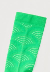 Zöld zokni, amely ismétlődő fehér mintával díszített, amely ívelt vonalakból áll, és Wi-Fi jelekre hasonlít, texturált, rugalmas anyagból készült.