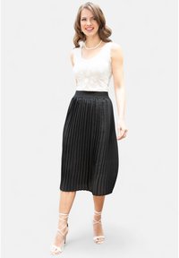 Gonna midi plissettata nera con finitura satinata, abbinata a un top senza maniche in pizzo bianco. Il modello indossa tacchi nudi con cinturini.