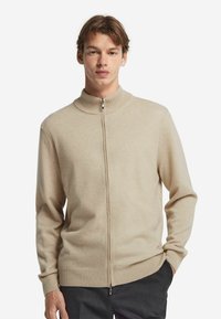 Beige zip-up tröja med hög krage, stickad textur och ribbade ärmar, bärs över svarta byxor. Har en silverfärgad dragkedja vid kragen.