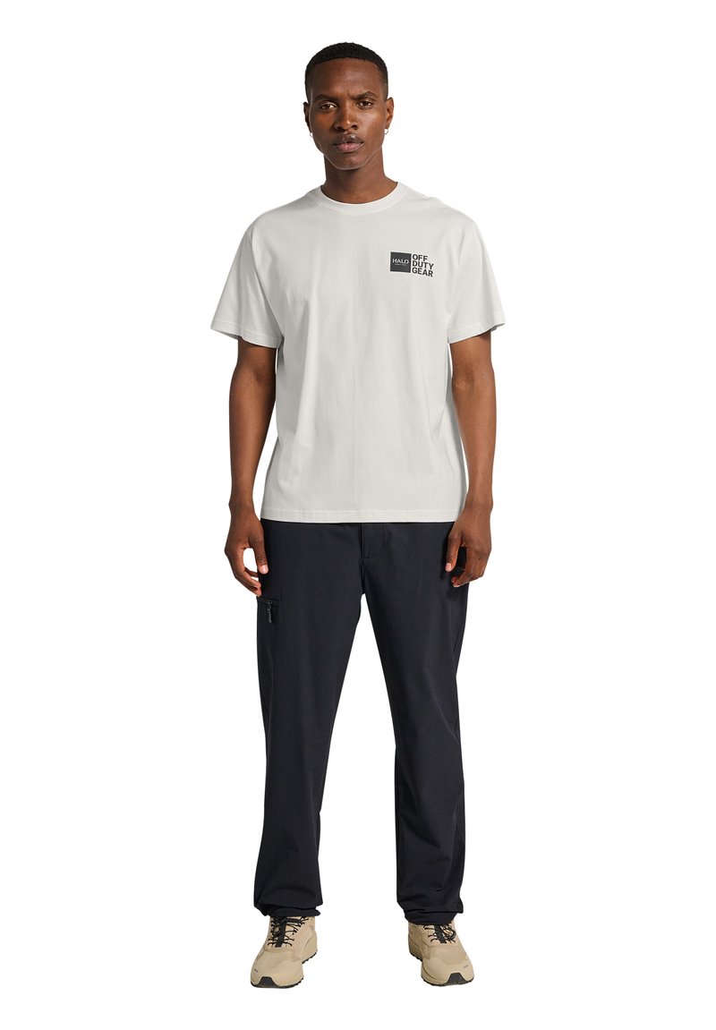 Man draagt een wit "HALO Off Duty Gear" T-shirt, zwarte broek met een ritszak en beige wandelschoenen, staand tegen een witte achtergrond.