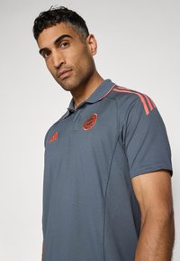 Grå polo skjorte med korte ærmer, prydet med lyse orange striber på skuldrene og et broderet logo på brystet. Tekstureret stof.