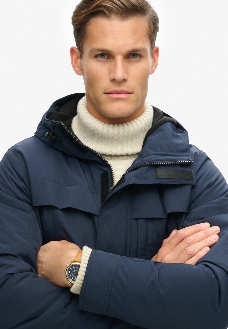 Marineblauwe donsjack met een capuchon, voorzien van een rits aan de voorkant en borstzakken met kleppen. Gelaagd met een crèmekleurige geribde turtlenecktrui.