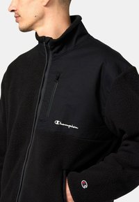 Schwarze Fleecejacke mit Reißverschlusstasche, kontrastierendem Nylonpanel und gesticktem Logo. Verfügt über einen hohen Kragen und strukturierter Stoff.
