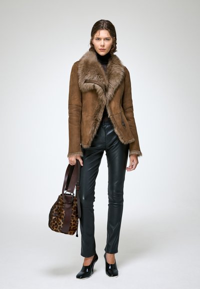 Femme portant une veste en suède marron doublée de fourrure, un pantalon en cuir noir, des talons hauts noirs et tenant un sac à main en fourrure à motif léopard.