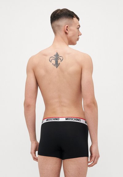 Mustat boxerit valkoisella ja punaisella vyötärönauhalla, jossa on "MOSCHINO"-logo; mallilla on näkyvissä taaksepäin tatuointi ja lyhyt hiustyyli.
