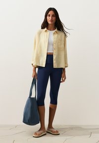 Femme portant une chemise rayée jaune par-dessus un crop top blanc, des leggings capri bleu marine, des sandales beiges, et tenant un grand sac cabas bleu.
