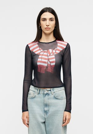 Γυναίκα με μακριά σκούρα μαλλιά φοράει διάφανο μαύρο μακρυμάνικο τοπ με γραφικό φουλάρι με κόκκινες και λευκές ρίγες, συνδυασμένο με ανοιχτό μπλε τζιν παντελόνι.