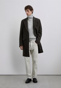 Abrigo de lana oscuro sobre un suéter de cuello alto gris claro, combinado con pantalones color crema y zapatos negros. Diseño sencillo, corte entallado.
