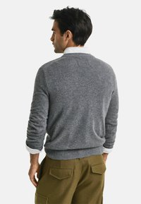 Un pull gris avec un ourlet et des poignets côtelés, porté par-dessus une chemise blanche à col. Le modèle est de dos, mettant en valeur le design et la coupe du dos.