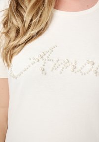T-shirt blanc avec des embellissements en forme de perles surélevées disposées en un motif décoratif sur le devant. Texture en coton lisse.