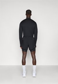 Schwarzes Langarmoberteil und schwarze Shorts mit einer inneren Schicht, kombiniert mit weißen Socken und grauen Sportschuhen; minimales Design und Texturen.