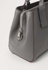 Sac à main en cuir gris texturé avec double fermeture éclair argentée et finition marron foncé, avec une poignée courte et un design en couches.