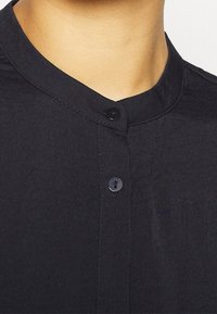 Sort knap-up beklædning med mandarin krave, lavet af en glat, struktureret stof og en enkelt sort knap lukning ved halsen.